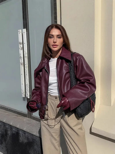 woman wearing maroon faux leather bomber jacket with zipper and beige pants holding sunglasses outdoors