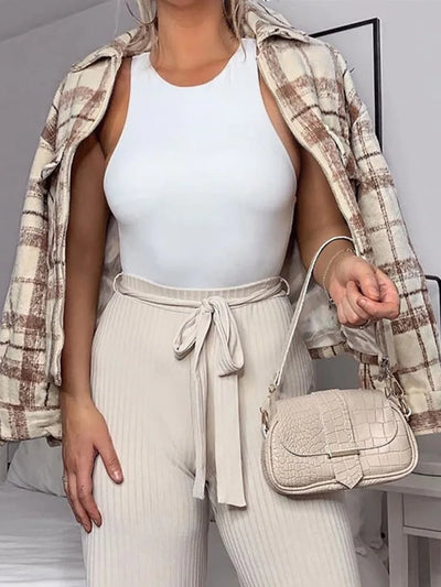 Woman wearing white sleeveless slim-fit bodysuit paired with beige high-waisted pants and plaid jacket, holding beige handbag